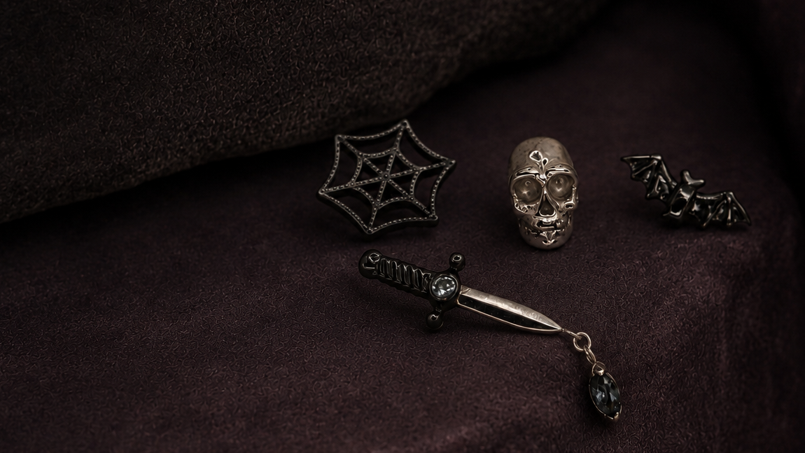 A gothic-themed jewelry set displayed on a dark brown velvet surface, featuring a silver skull ring, a black spiderweb brooch, a silver dagger with a black teardrop charm, and a black bat charm.