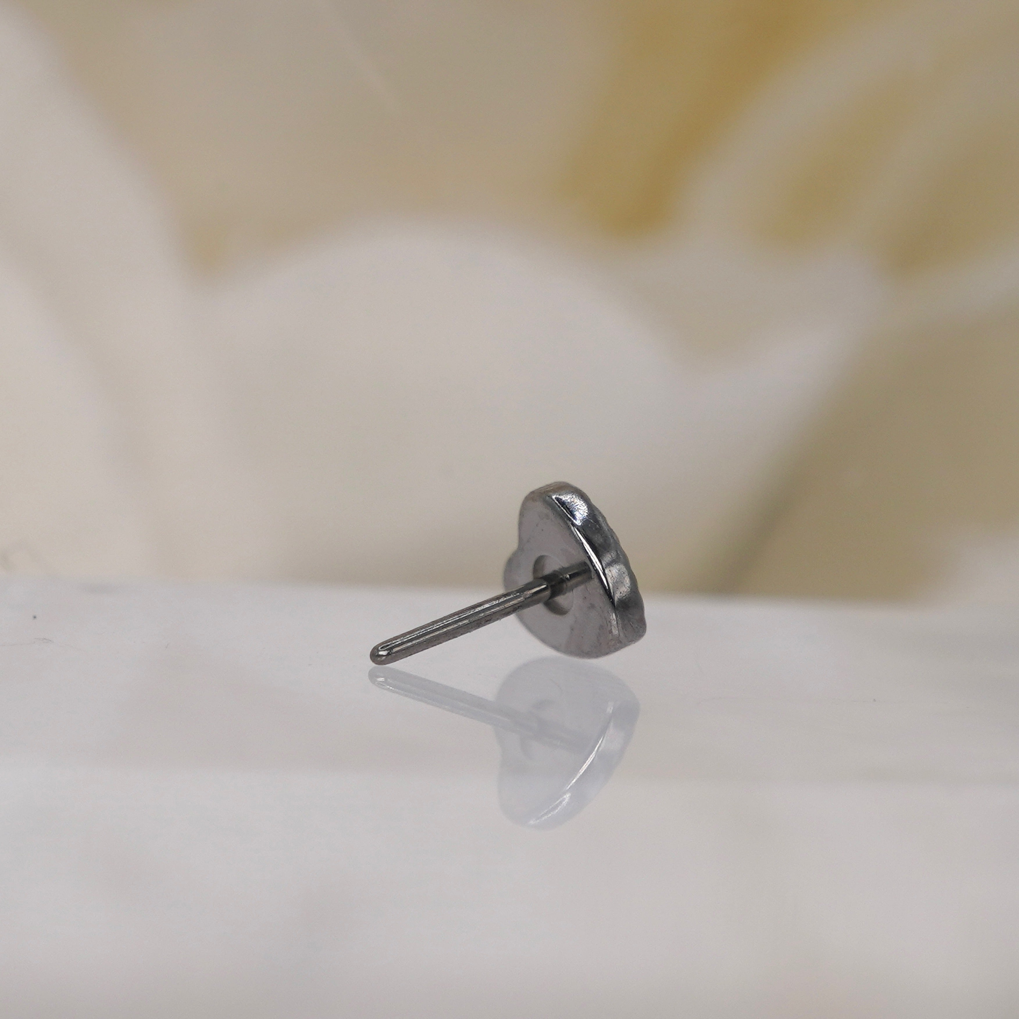 Threadless Implant Grade Titanium Laser Heart by Junipurr, shown on a reflective white surface with a soft, out-of-focus cream background.