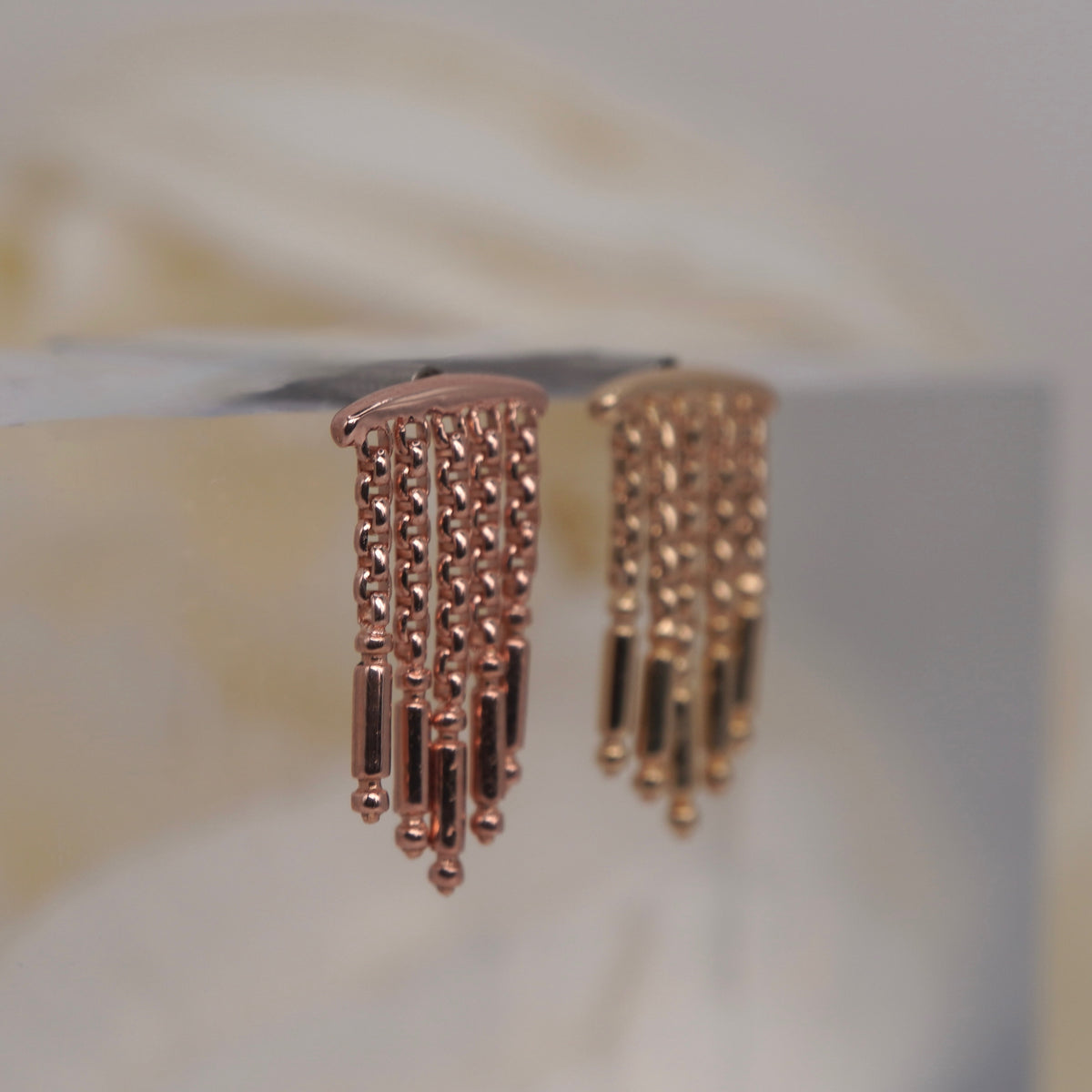 Symphony Hidden Helix 16g leroi dangling earrings with multiple chains of small beads and cylindrical links, displayed on a clear stand against a soft beige background side view.