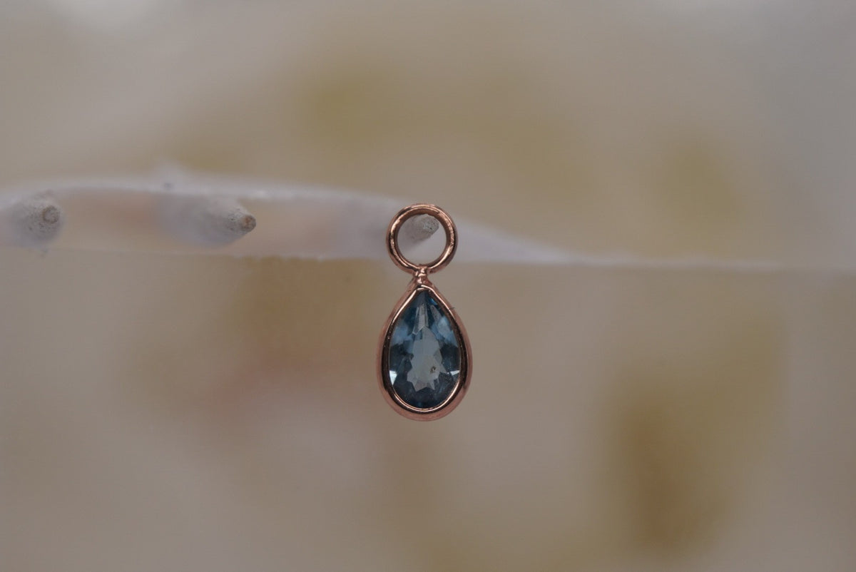 Front view of Buddha Jewelry’s “Concorde Teardrop” in 14k rose gold, featuring a faceted London Blue Topaz drop in a smooth gold setting.