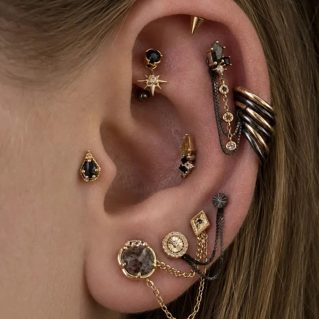 A close-up of a person's ear adorned with multiple gold-toned and black gothic piercings, including ear cuffs, studs, and a chain-link cartilage piercing.