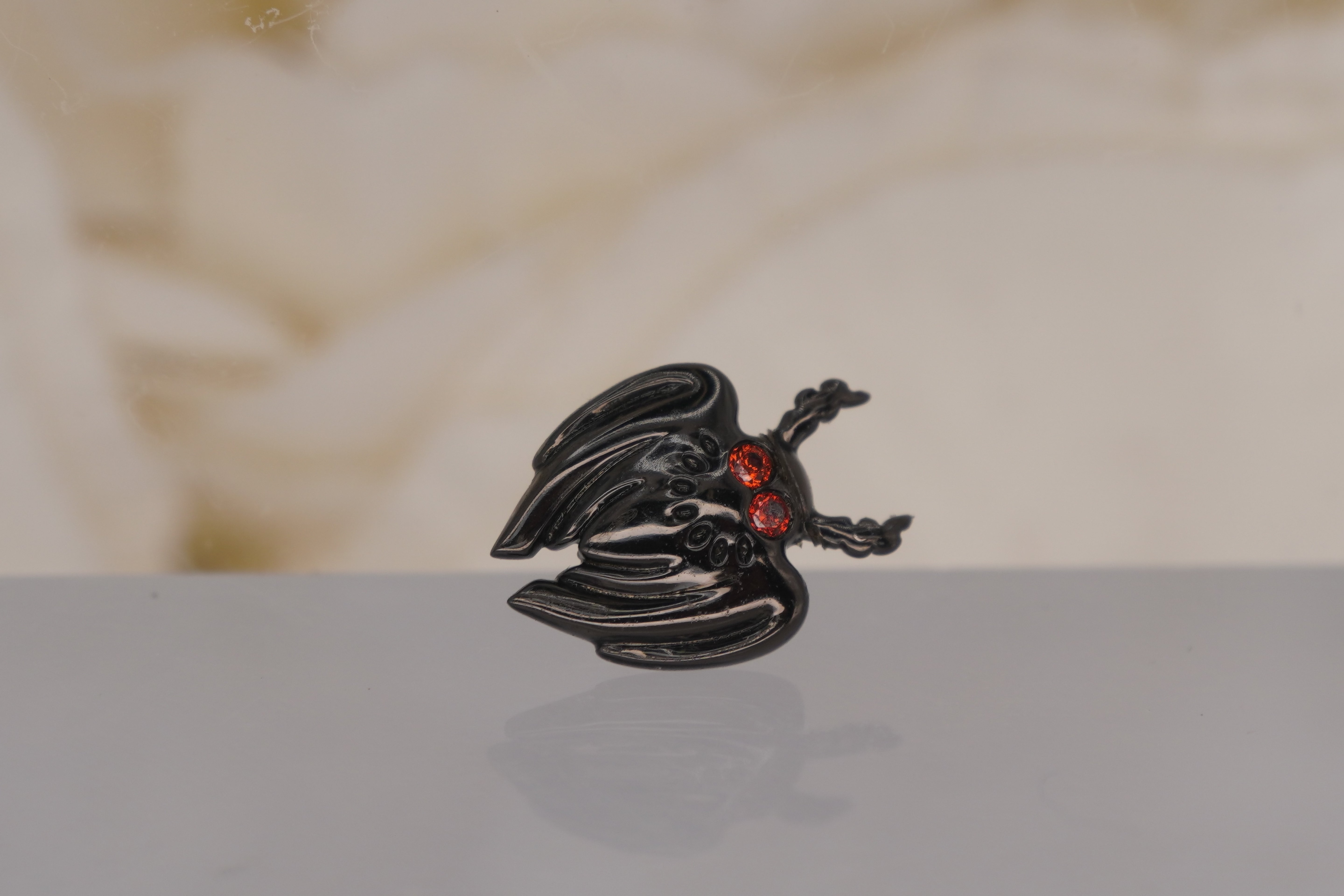 Junipurr 14k Gold “Scene Mothman” Threadless End in Black Rhodium garnet eyes resting on a reflective white surface with a soft beige fabric background.