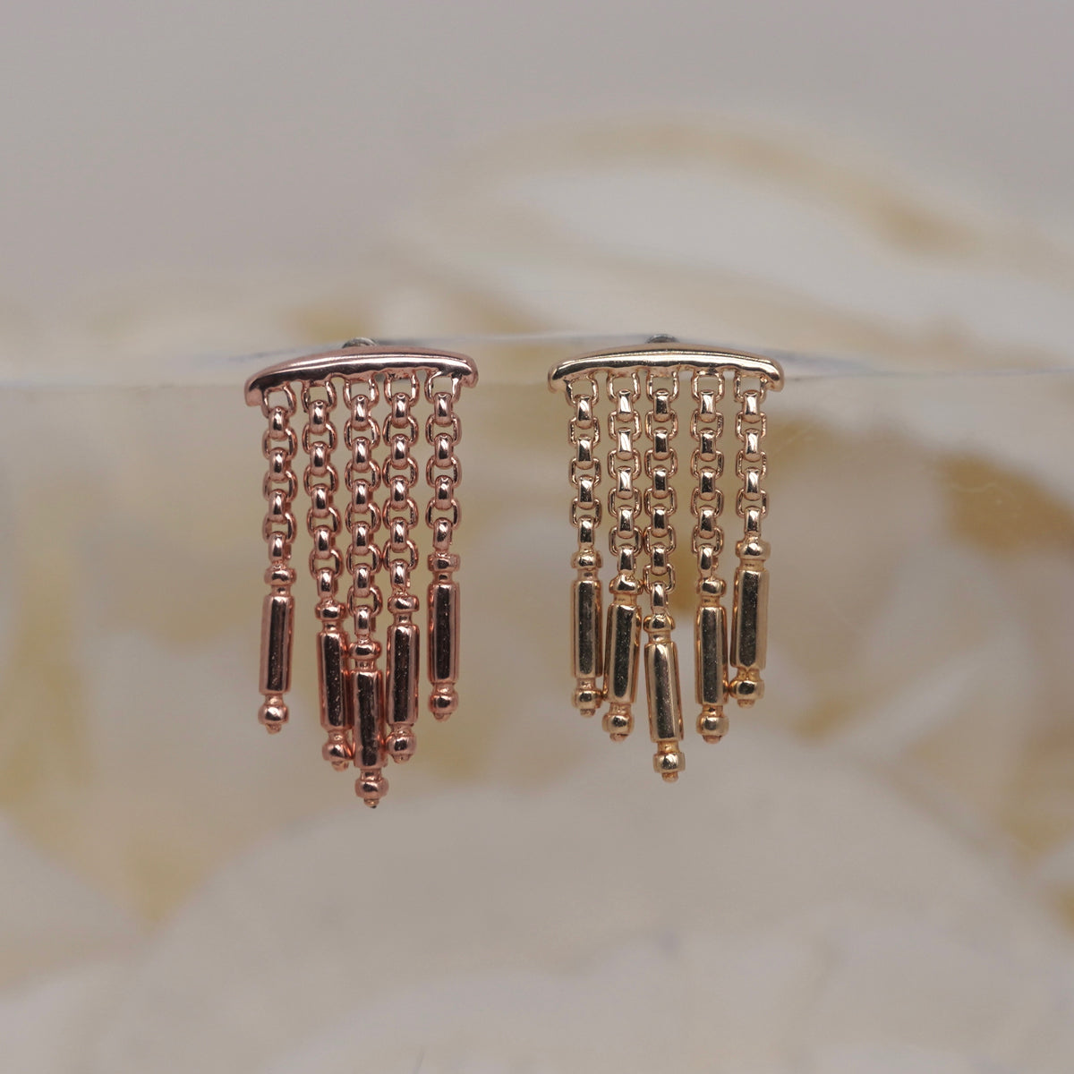 Symphony Hidden Helix 16g leroi yellow gold rose gold dangling earrings with multiple chains of small beads and cylindrical links, displayed on a clear stand against a soft beige background.