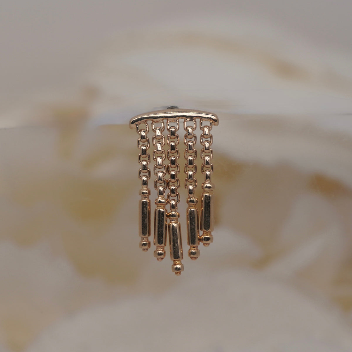 Symphony Hidden Helix 16g leroi yellow gold dangling earrings with multiple chains of small beads and cylindrical links, displayed on a clear stand against a soft beige background.