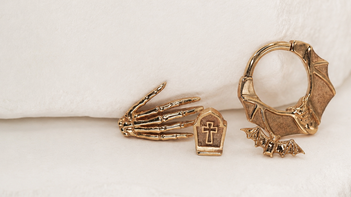 Gold-toned gothic jewelry set featuring a skeletal hand, a tombstone with a cross, and a bat pendant, arranged on a soft white textured fabric background.