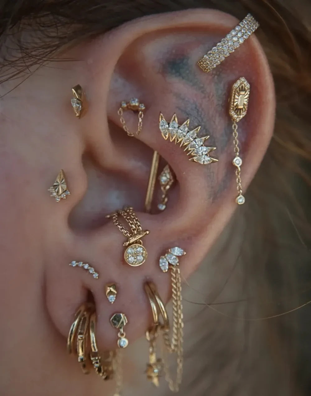 A close-up of a person's ear adorned with multiple gold and silver cartilage piercings and studs, including chain earrings, hoop earrings, and crystal-studded jewelry.