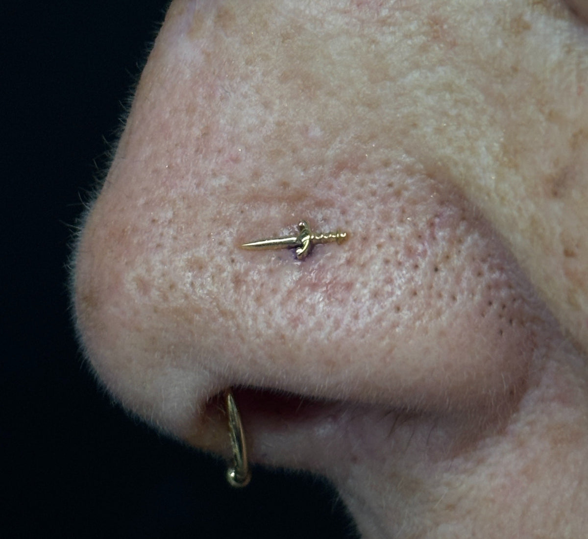 Buddha Jewelry’s “Blade” worn in a nose piercing, showing its elongated sword shape and sharp, polished details.